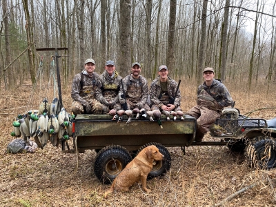 Five men in camouflage clothes sitting on a trailer with ducks and duck decoys and a 3-legged tan dog sitting in front